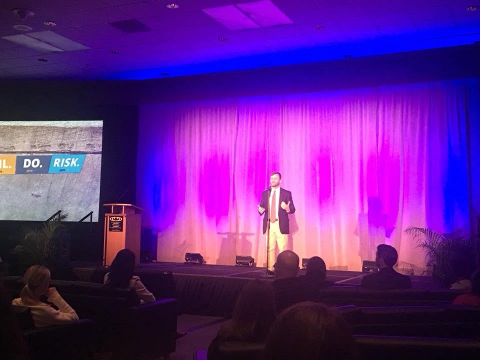 David Cutler speaking on a conference stage with purple lighting and an audience in the foreground.